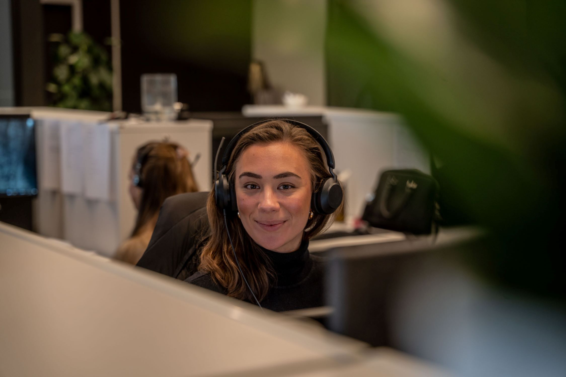 Josephine sitter ved skrivebordet på kontoret hos Byggfakta, iført headset og smilende mens hun ser inn i kameraet. Man ser kontormiljøet i bakgrunnen.